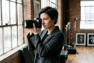 Photographe professionnelle tenant un appareil photo reflex dans un studio avec murs en briques apparentes, illustrant le choix de carrière entre photographie et vidéographie